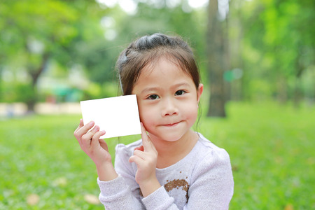 Adorable little Asian child girl showing up a blank white paper in the green gardenの写真素材