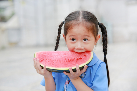 Smiling little Asian child girl in school uniform enjoy eating watermelon outdoorsの写真素材