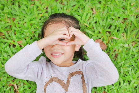 Portrait of smiling cute little Asian child girl lying on green lawn with making her hands for heart sign on eyeの写真素材