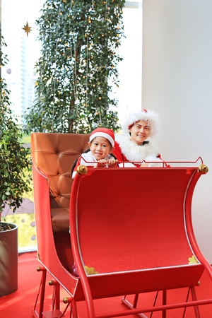 Happy little child girl and her mother in santa costume dress sitting on red sledge christmas background.  Merry Christmas and happy new year.の写真素材