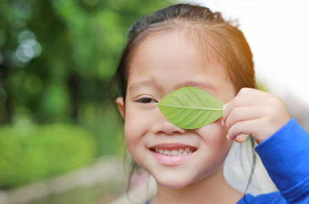 Adorable little Asian child girl holding a green leaf closing left eye in green garden background.の写真素材