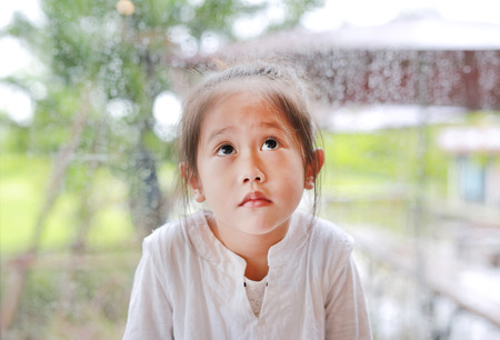 Little Asian child girl gesture with peaceful face and looking up against glass window with water drop at rainy day.の写真素材