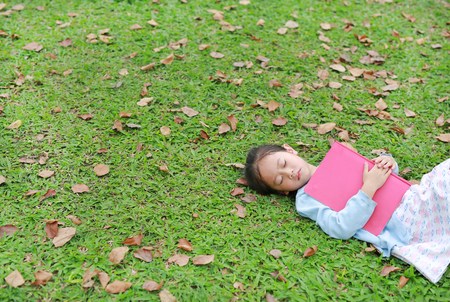 Little girl resting with book lying on green grass with dried leaves in the summer garden.の写真素材
