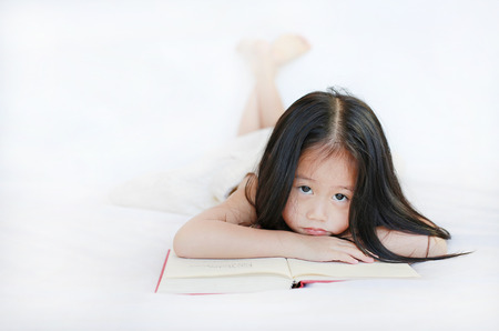 Adorable little Asian girl with hardcover book lying on bed and looking camera over white background.の写真素材