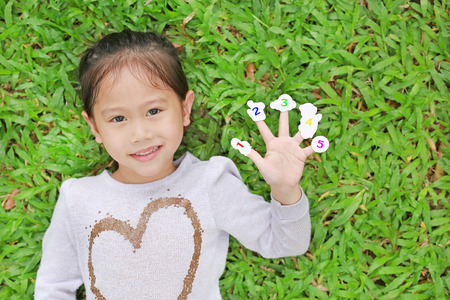 Smiling little Asian child girl lying on green grass lawn with showing white stickers with number...