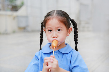 Adorable little Asian child girl in school uniform enjoy eating sausage.の写真素材