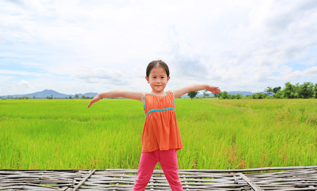 Asian kid girl stretch arms and relaxed at the young green paddy fields with mountain and cloud sky.の写真素材
