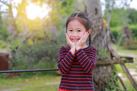 smiling little Asian kid girl with touching her cheek looking straight at camera in the nature garden.の写真素材