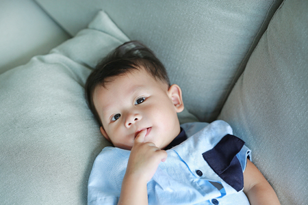 Adorable little Asian baby boy sucking finger in mouth lying on gray sofa.