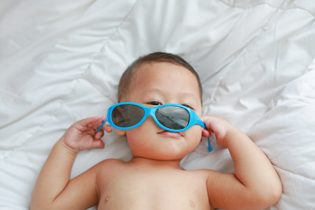 Portrait little Asian baby boy with sunglasses lying on white blanket on bed. Above view.の写真素材