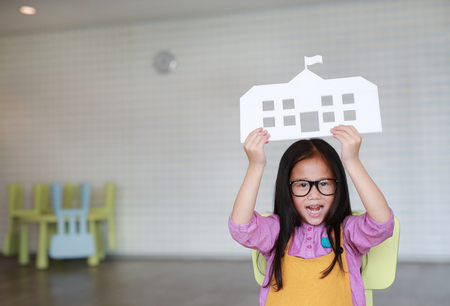 Happy little Asian child girl in pink-yellow dungarees holding mock-up paper school sitting on kid chair in classroom with looking straight at camera. Education and back to school concept.の写真素材