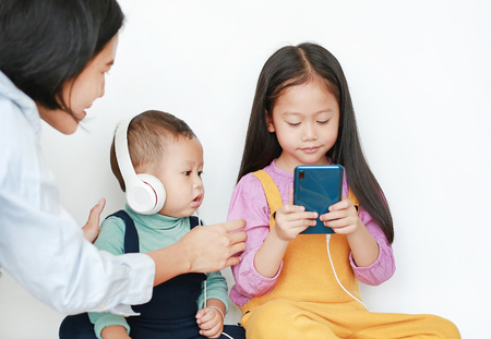 Mother taking Asian older sister and little brother sharing to enjoys listening music with headphones by smartphone isolated over white background. Sharing concept.の写真素材