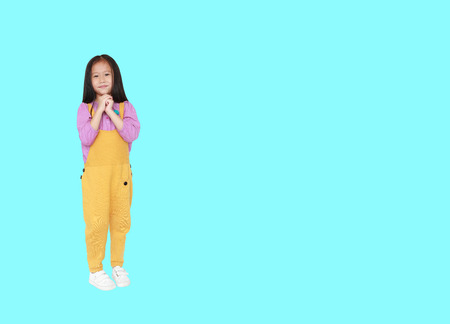Smiling little Asian child girl in pink-yellow dungarees expression hands implore isolated over cyan...