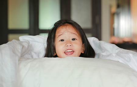 Smiling little girl lying under a white blanket on the bed.の写真素材