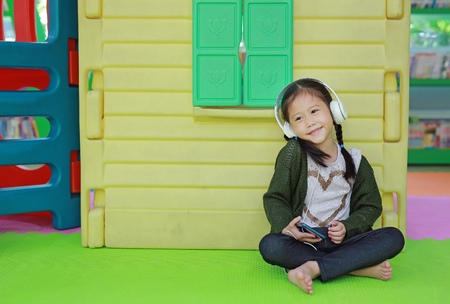 Cute little Asian child girl listening music by headphone near toy playhouse in playground.の写真素材