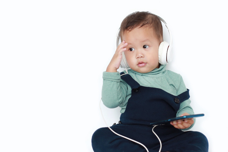 Adorable Asian little baby boy enjoys listening music with headphones by smartphone isolated over white background.の写真素材