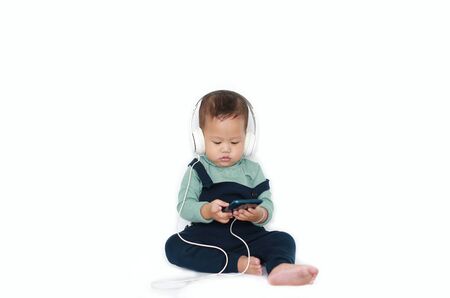 Adorable Asian little baby boy enjoys listening music with headphones by smartphone isolated over white background.の写真素材