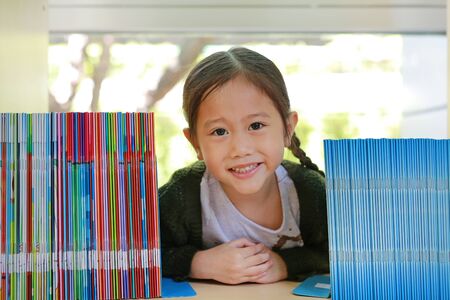 Smiling little Asian child girl lying on bookshelf at library. Children creativity and imagination concept.の写真素材