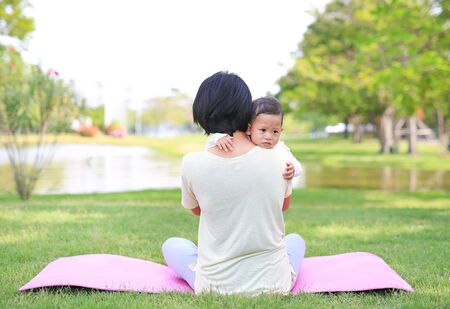 Asian mom carrying son and sitting on green lawn in nature park.の写真素材