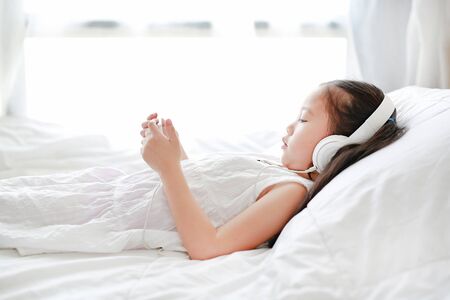 Portrait little Asian girl using headphones listen music by smartphone while lying on bed at home.