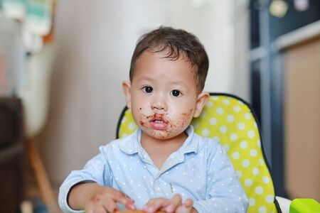 Asian little baby boy sitting on children chair indoor eating bread with Stuffed Chocolate-filled dessert and Stained around her mouth.の写真素材