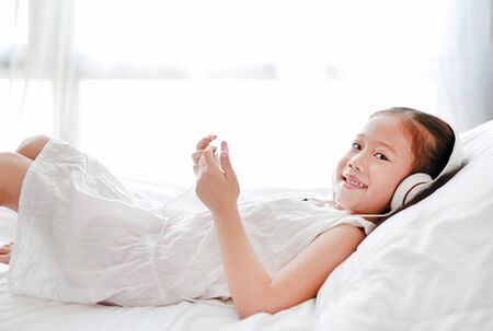 Happy little Asian girl using headphones listen music by smartphone smiling and looking camera while lying on bed at home.の写真素材