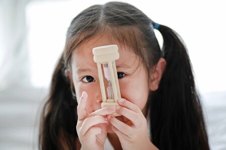 Close up little Asian girl holding sandglass in hand with looking through camera. Waiting times with hourglass.の写真素材