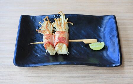 Bacon and Enoki Mushroom roll stick served with lemon on black plate on wood table. Japanese cuisine...