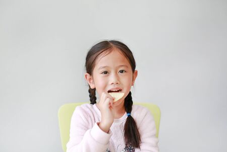Adorable happy little Asian child girl eating crispy potato chips on white background. Kid enjoy...