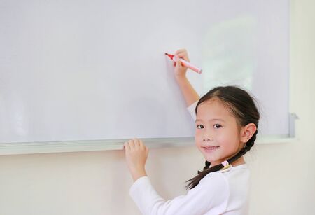 Portrait of happy little Asian child girl or Schoolgirl writing something on whiteboard with a marker and looking at camera in the classroom. White board with copy space for text.の写真素材