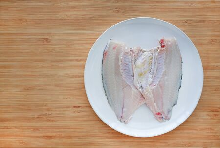 Raw tilapia fish fillet on white plate against wooden board background with copy space.