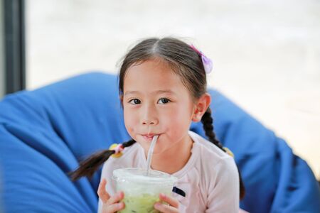 Happy little Asian child girl drinking iced Matcha Green Tea lying on sofa at cafe.の写真素材
