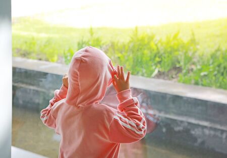 Child in pink tracksuit with hood on head at glass window rear view looking out to green lawn outdoor.の写真素材