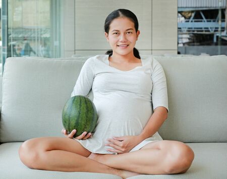 Pregnant woman sitting on sofa and holding watermelonの写真素材