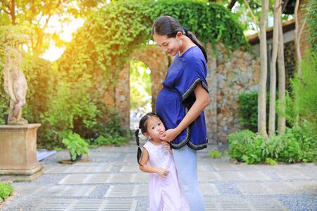 Beautiful little girl hugging a pregnant and kissing mother's belly in the garden.の写真素材