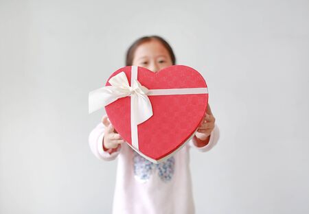 Adorable little Asian child girl showing red heart gift box on white background. Kid giving red heart gift box for you. Concept of love.の写真素材