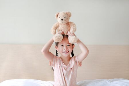 Happy Asian little girl Picking up brown teddy bears on her head while sitting on the bed at home.の写真素材