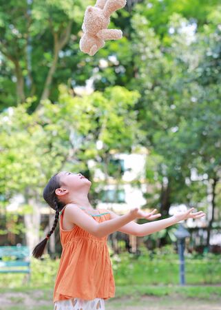 Portrait of happy little Asian child in green garden with throwing up teddy bear doll floating on air. Smiling kid girl playing in summer park.の写真素材