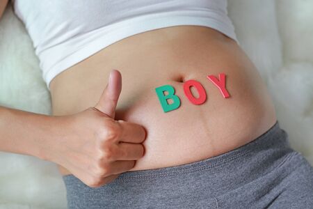 Close up Pregnant woman sitting on soft sofa and touching her belly with sign BOY in front of her belly. Showing thumbs up sign.の写真素材