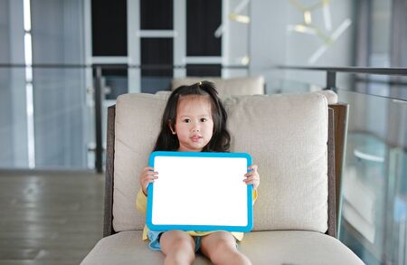 Cute little child girl holding empty white blackboard on fabric sofa in library room.