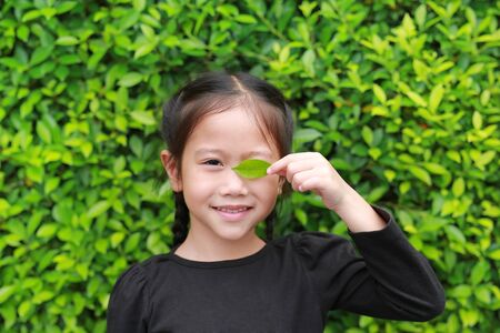 Portrait of smiling little Asian child girl holding a green leaf closing right eye in green garden background.の写真素材