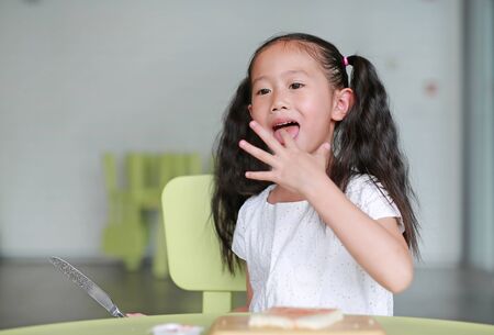 Portrait happy little child girl cooking at class room. Kid taste strawberry jam while cooking. Children are using knife slicing and apply jam on bread on chopping wood board.の写真素材