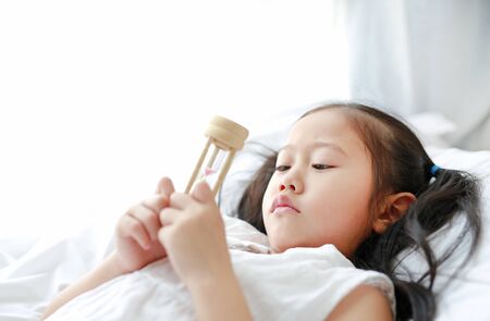 Portrait of little Asian girl looking at hourglass in hand lying on bed at home. Waiting times with sandglass. Close-up shot.の写真素材