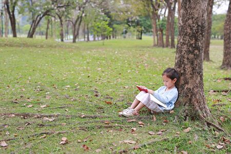 Adorable little girl reading a book sitting under a tree outdoor garden.の写真素材