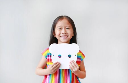Happy little child girl holding blank white animal paper mask on her chest on white background. Idea and concept for kid dressed up playing animal face.の写真素材