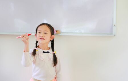 Portrait of happy little Asian child girl or Schoolgirl holding a marker against whiteboard in the...