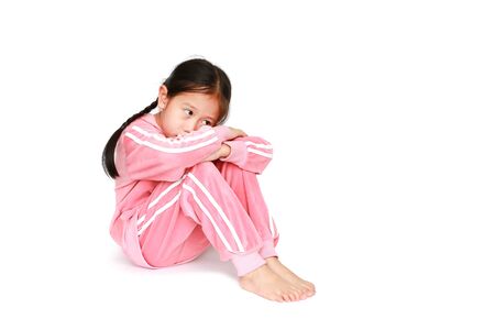 Sad little Asian child girl in pink tracksuit or sport cloth sitting and hugging knees over white background with looking out.の写真素材