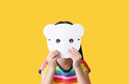 Little child girl holding blank white animal paper mask fronting her face isolated on yellow background. Idea and concept for kid dressed up playing animal face.の写真素材