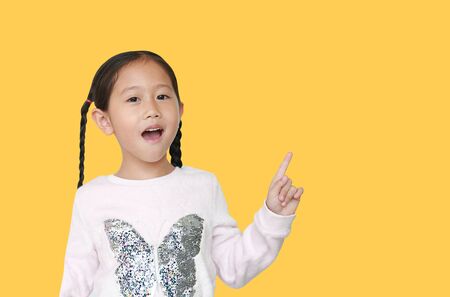 Happy Asian little child girl pointing forefinger up isolated over yellow background with copy space. Cheerful emotion concept.の写真素材