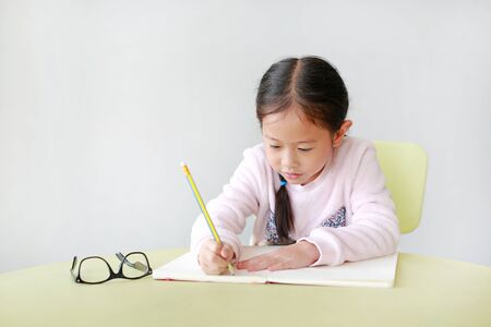 Portrait of smiling little Asian girl writes in a book or notebook with pencil on table in classroom...